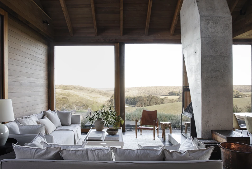 Contemporary living room interior features neutral sofas, a concrete fireplace, a leather chair, and a landscape view through large windows.