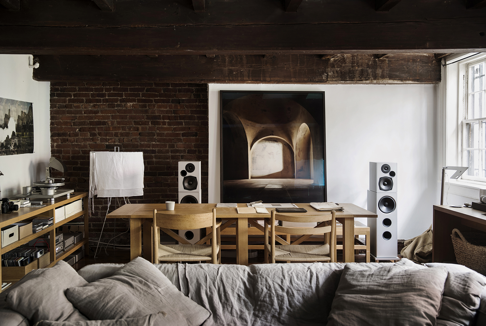 Living room interior with a linen sofa, wooden table and chairs, speakers, and a large artwork in a natural light setting.