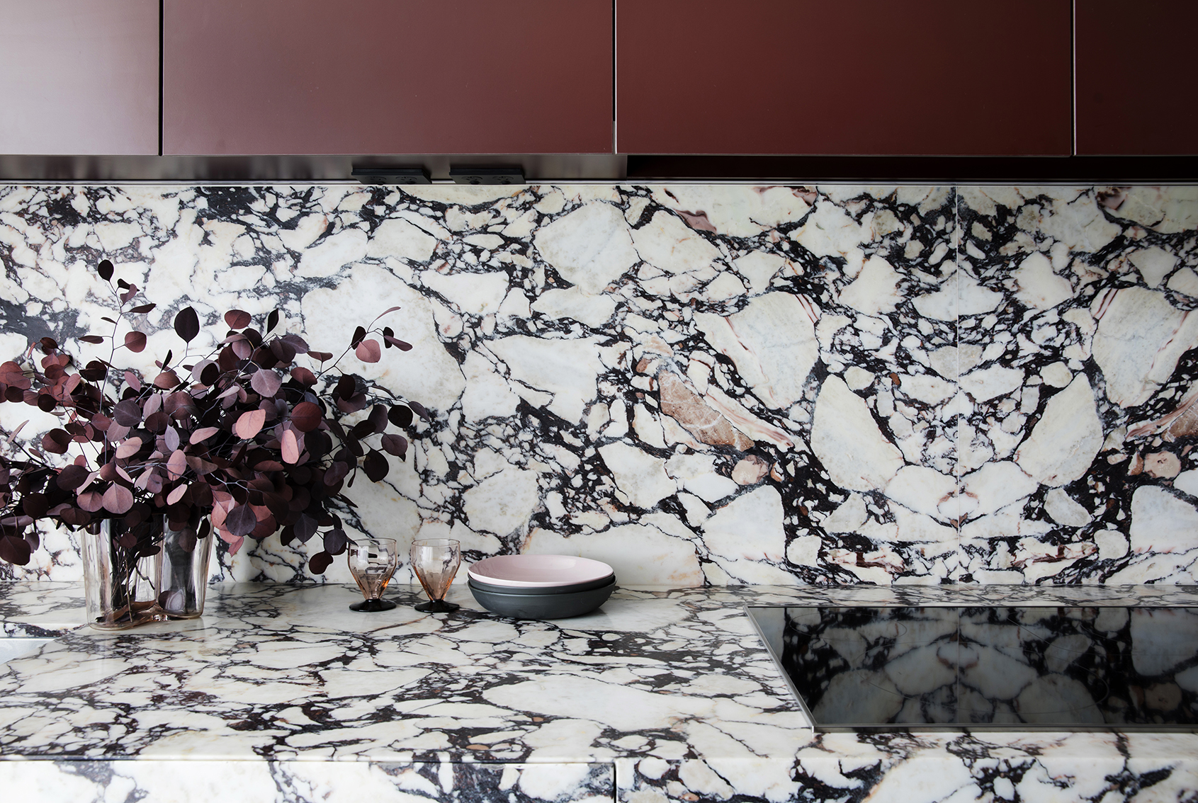 Modern kitchen interior featuring a marble countertop and backsplash, burgundy cabinetry, a vase with burgundy leaves, and tableware.