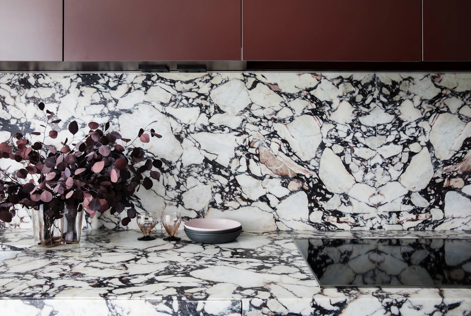 Modern kitchen interior featuring a marble countertop and backsplash, burgundy cabinetry, a vase with burgundy leaves, and tableware.