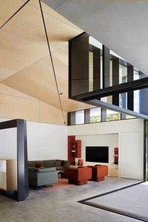 Modern living room interior with angular architecture, green sofa, rust-colored armchairs, a black TV, and large windows with natural light.
