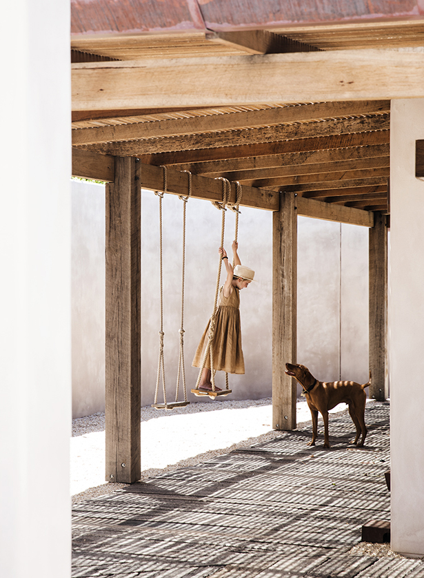 Child in a brown dress swinging on a wooden swing under a wood-beamed roof, with a dog watching on a sunny day.