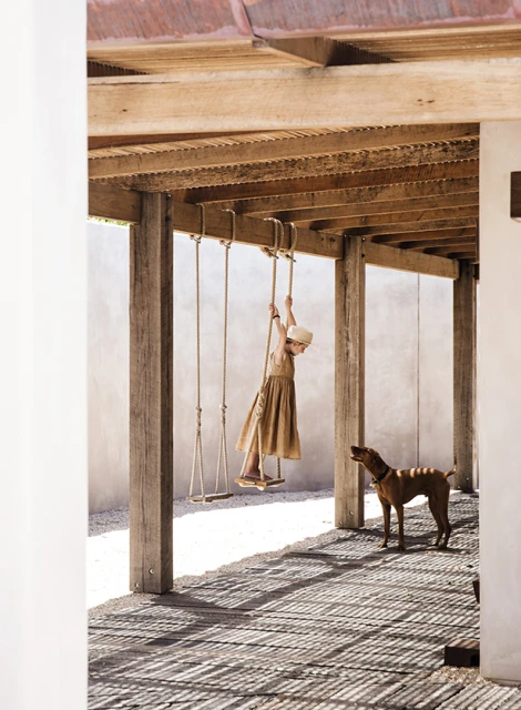Child in a brown dress swinging on a wooden swing under a wood-beamed roof, with a dog watching on a sunny day.