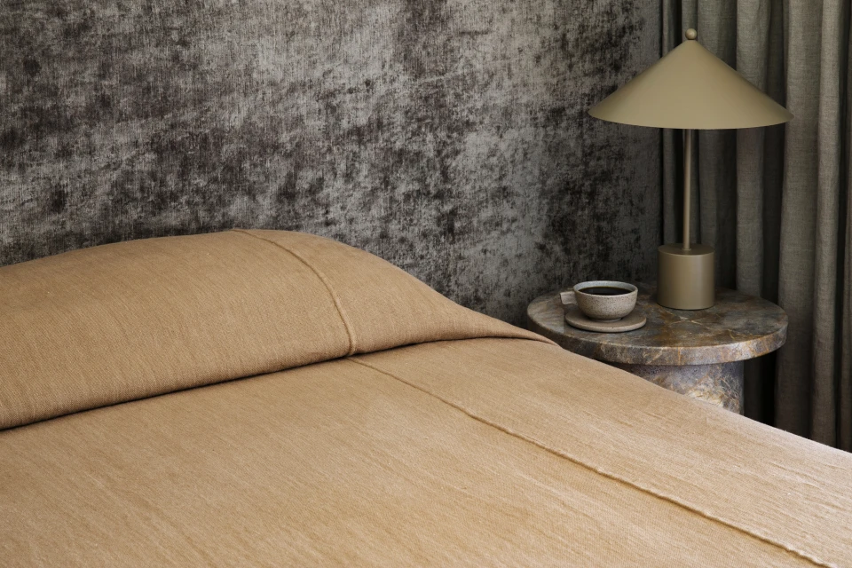Bedroom interior featuring a tan bed with a matching headboard, table lamp, round side table, coffee cup, and a textured wall.