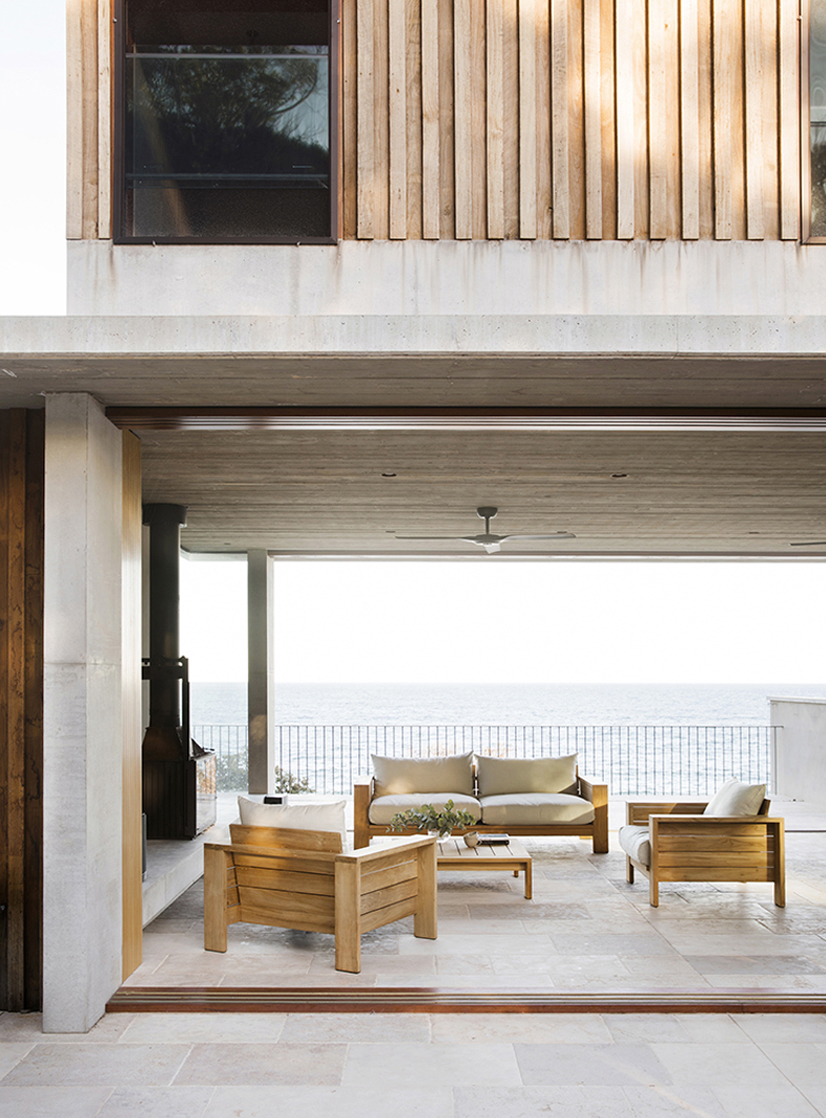 Exterior view of modern architectural design featuring an open-air patio with wooden furniture overlooking the ocean on a bright day.