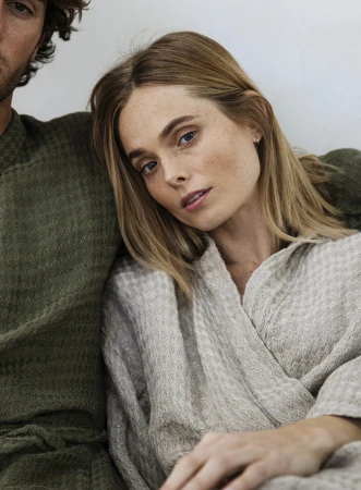 Close-up of a blonde woman in a robe leaning against a man in knitwear, showcasing a cozy, intimate setting and focus on textures and tones.
