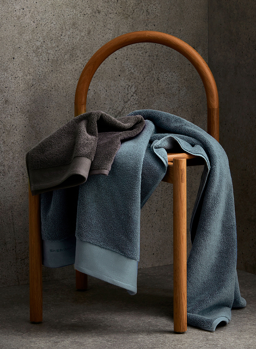 Stacked towels on a wooden chair against a textured grey wall. Blue and grey towels are prominently displayed in an interior setting.