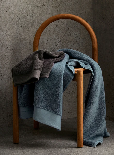 Stacked towels on a wooden chair against a textured grey wall. Blue and grey towels are prominently displayed in an interior setting.