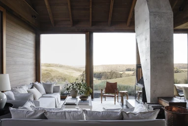 Modern living room interior with a large white sofa, concrete fireplace, and large glass windows overlooking a rolling landscape.