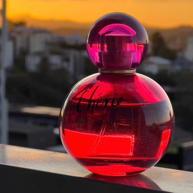 Close-up of a red Kate Spade Cherie perfume bottle against a blurred background of city buildings and a sunset sky.