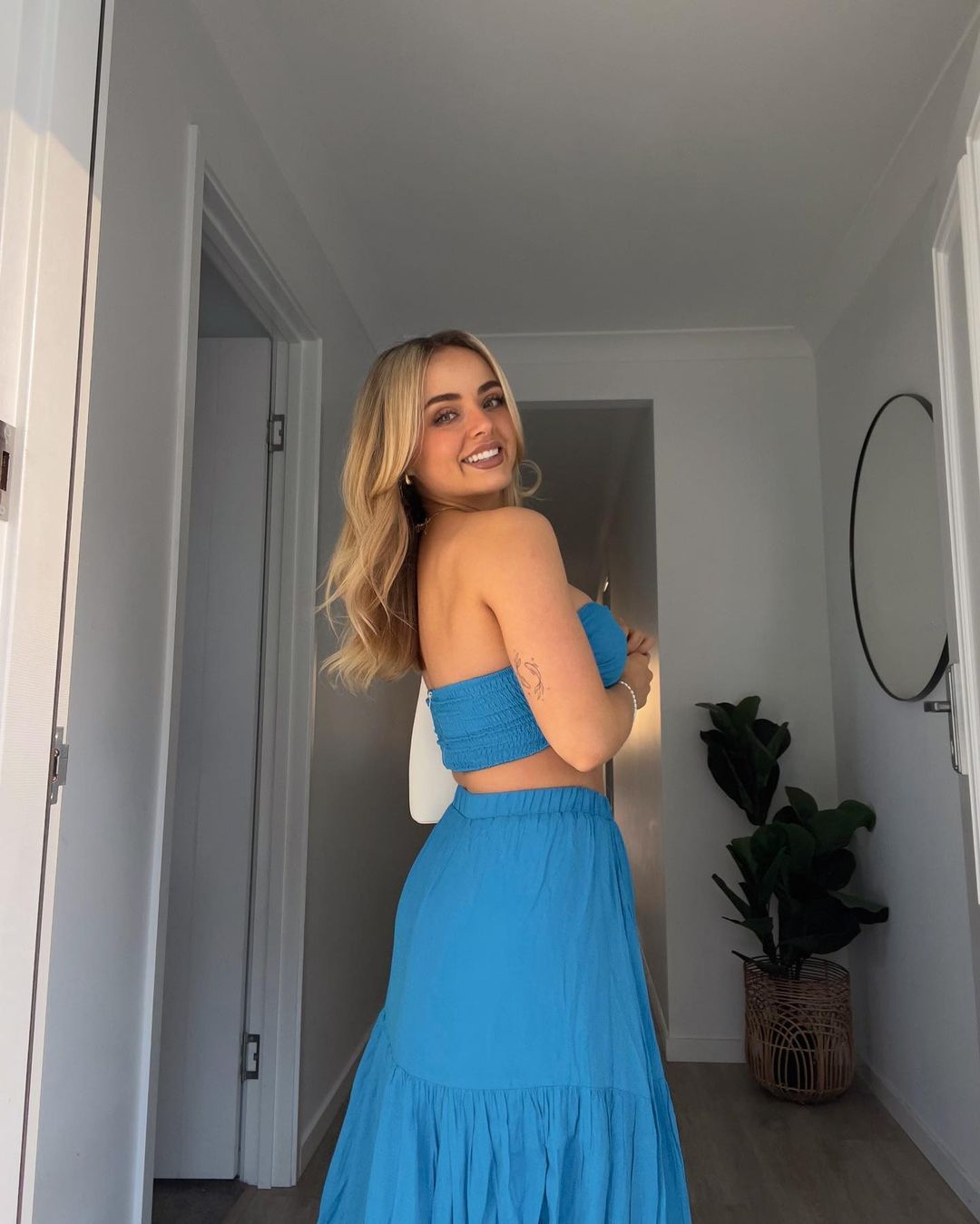 Blonde woman in a blue two-piece outfit poses in an indoor hallway, with a white door and a plant on a wicker basket.
