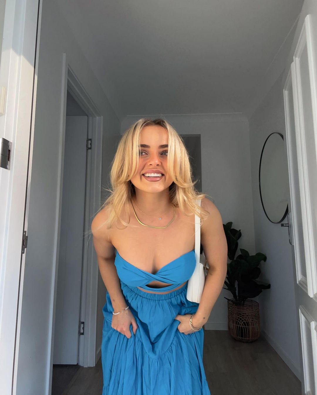 Blonde woman in a turquoise sundress smiles in an indoor setting. She is posing and carrying a small white purse over her shoulder.