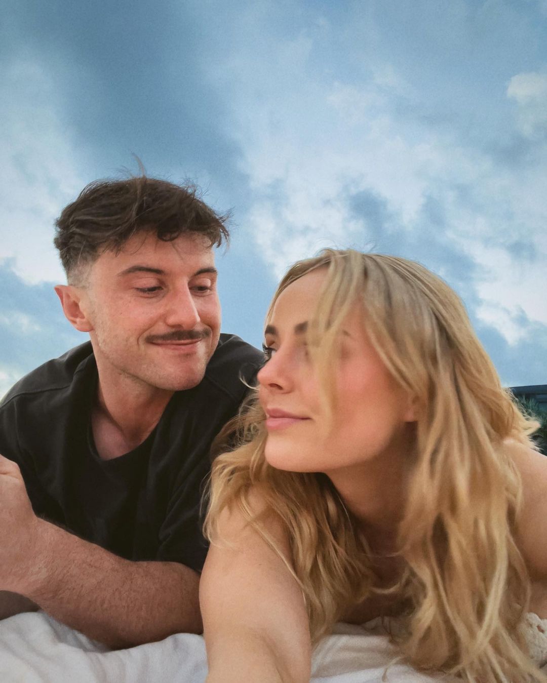 A tanned couple lies on their sides on a white blanket outside against a cloudy sky. The man looks at the woman, who gazes into the distance.