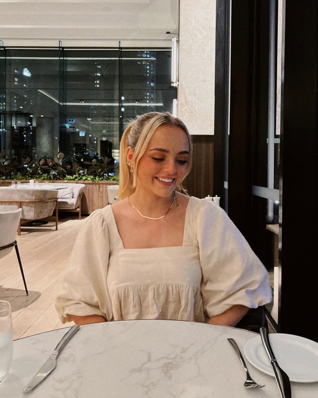 Blonde woman wearing a cream square-neck top seated at a round marble table in a restaurant by Conagh Kathleen