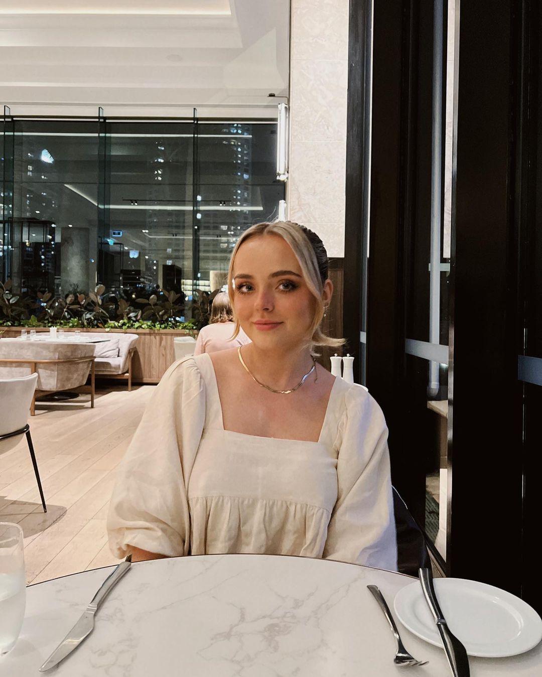 Blonde woman in neutral dress sits at a marble table in a restaurant with a large window, creating an inviting indoor portrait.