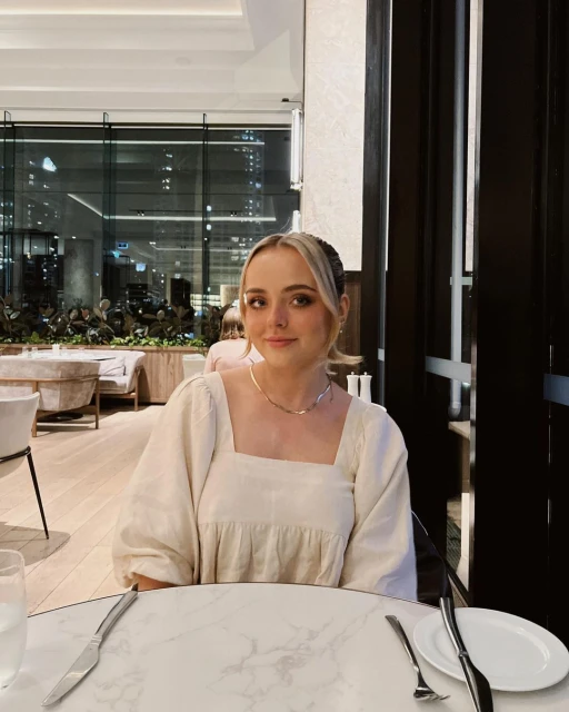 Blonde woman in neutral dress sits at a marble table in a restaurant with a large window, creating an inviting indoor portrait.