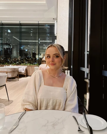 Blonde woman in neutral dress sits at a marble table in a restaurant with a large window, creating an inviting indoor portrait.