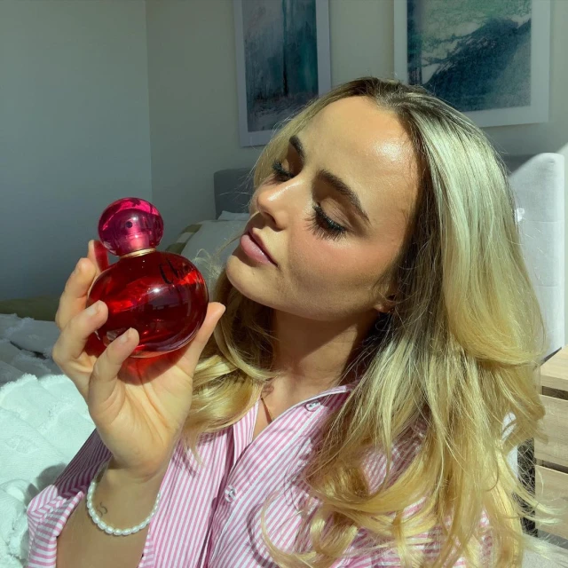 Blonde woman in pink shirt holding perfume bottle indoors, showcasing beauty product against a soft-focused bedroom backdrop.