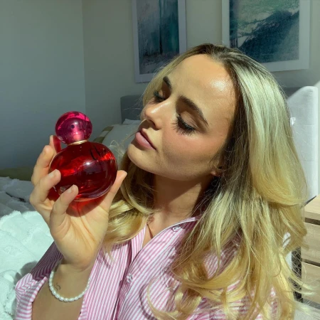 Blonde woman in pink shirt holding perfume bottle indoors, showcasing beauty product against a soft-focused bedroom backdrop.