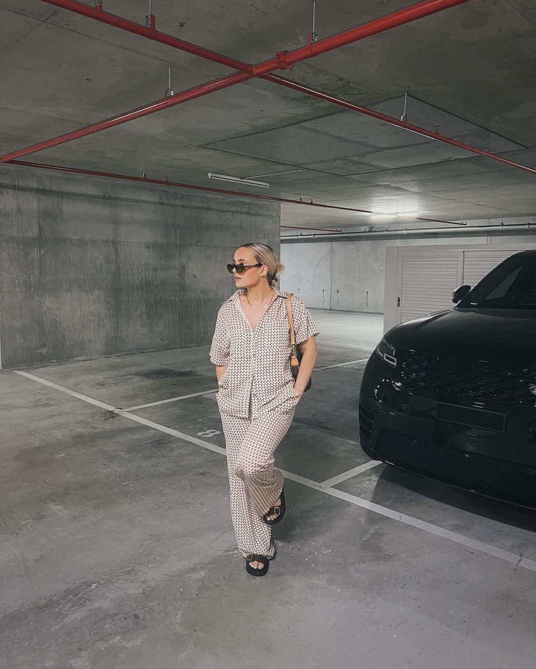 Woman in a checkered print outfit stands in a parking garage near a black Range Rover, wearing sunglasses and a shoulder bag. 