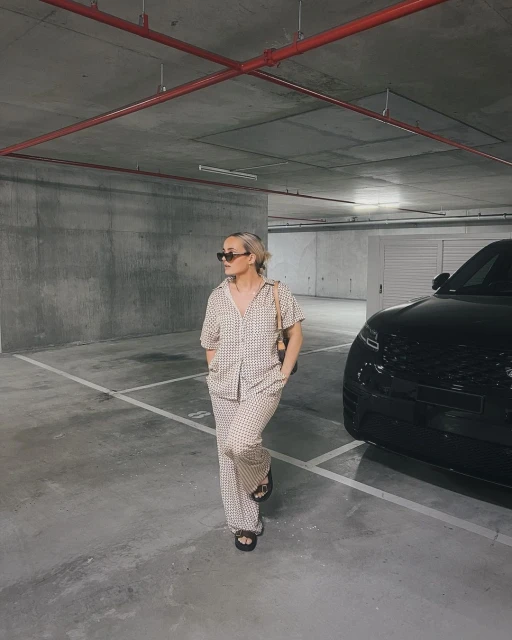 Woman in a checkered print outfit stands in a parking garage near a black Range Rover, wearing sunglasses and a shoulder bag. 