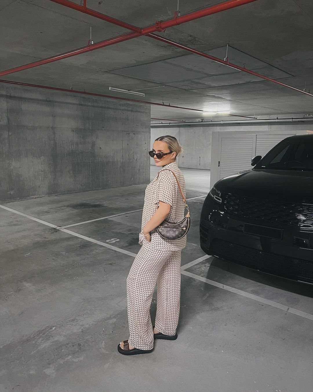 Woman in patterned shirt and pants stands in parking garage next to a black SUV, carrying a shoulder bag and wearing sunglasses.