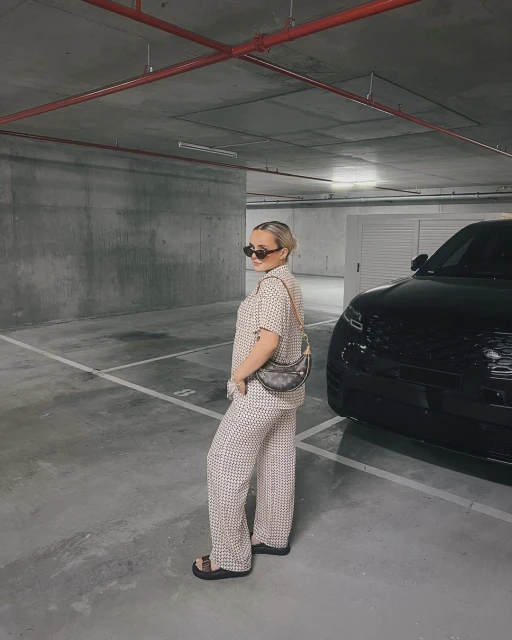 Woman in patterned shirt and pants stands in parking garage next to a black SUV, carrying a shoulder bag and wearing sunglasses.