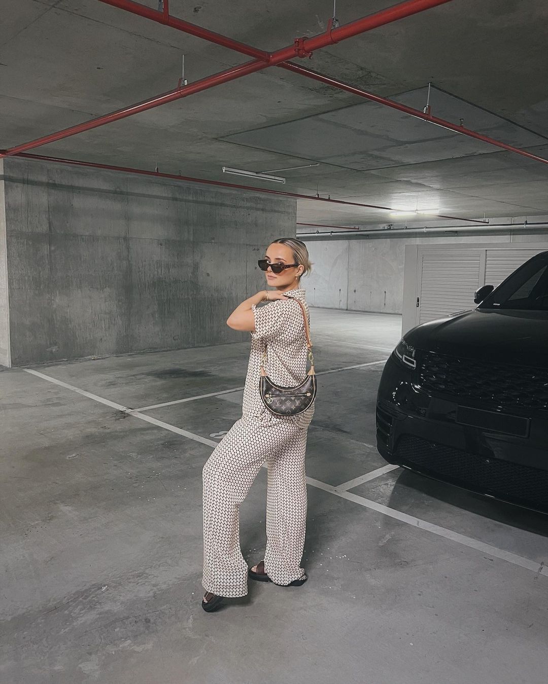 Stylish adult female poses in patterned pajama set in a parking garage with a black SUV and handbag. Cool, fashionable. 