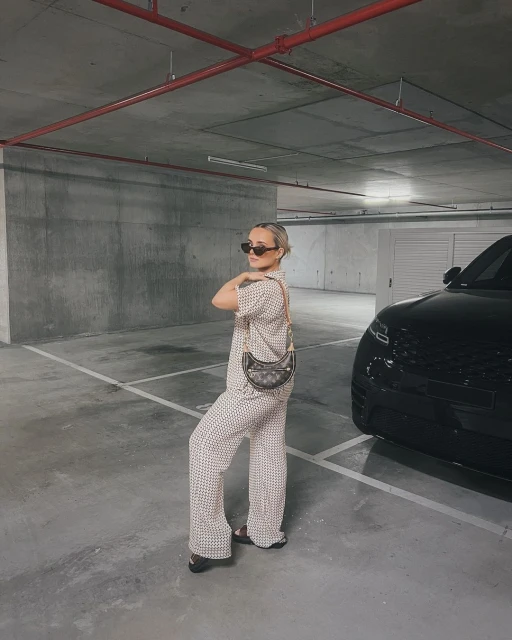 Stylish adult female poses in patterned pajama set in a parking garage with a black SUV and handbag. Cool, fashionable. 