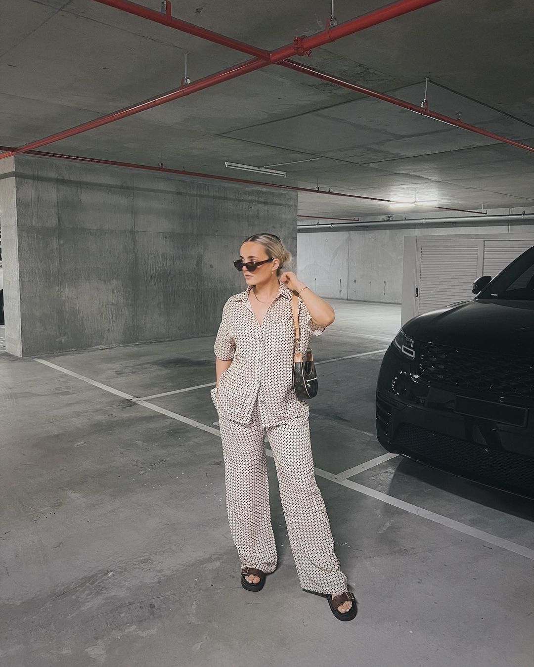 Woman in a patterned co-ord set poses in a parking garage with a handbag, sunglasses, and sandals, with a black car in the background.