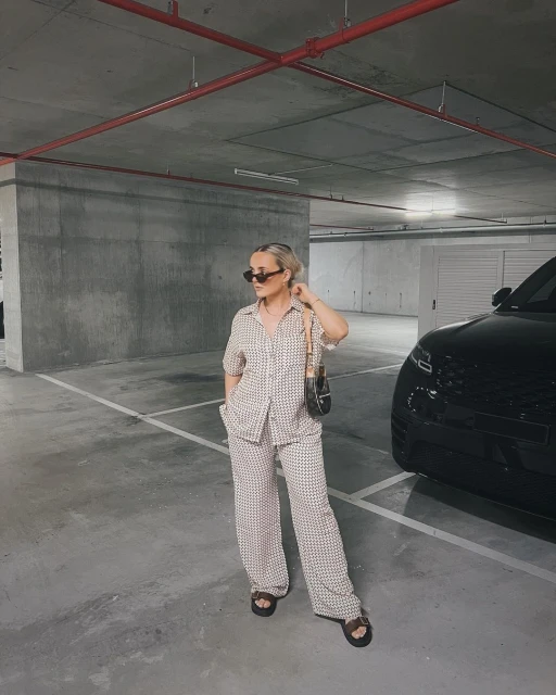Woman in a patterned co-ord set poses in a parking garage with a handbag, sunglasses, and sandals, with a black car in the background.