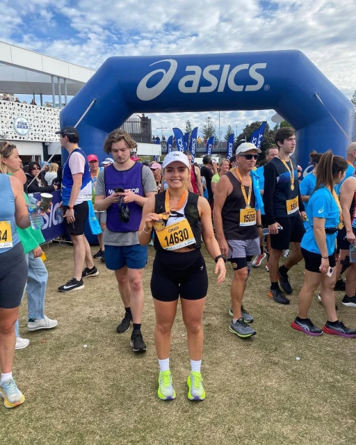 Female runner stands smiling after race, holding medal; outdoor, grassy field, Asics archway, crowd of people, sunny sky.