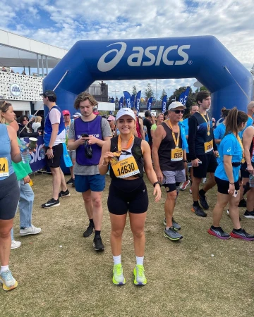 Female runner stands smiling after race, holding medal; outdoor, grassy field, Asics archway, crowd of people, sunny sky.