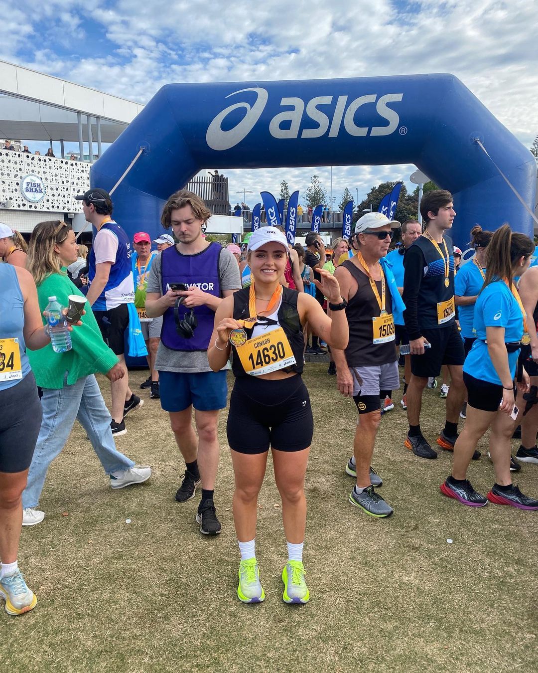 Female marathon runner stands with her race medal in front of an Asics archway with a large crowd of runners at the event.