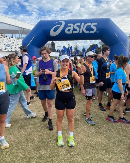 Female marathon runner stands with her race medal in front of an Asics archway with a large crowd of runners at the event.