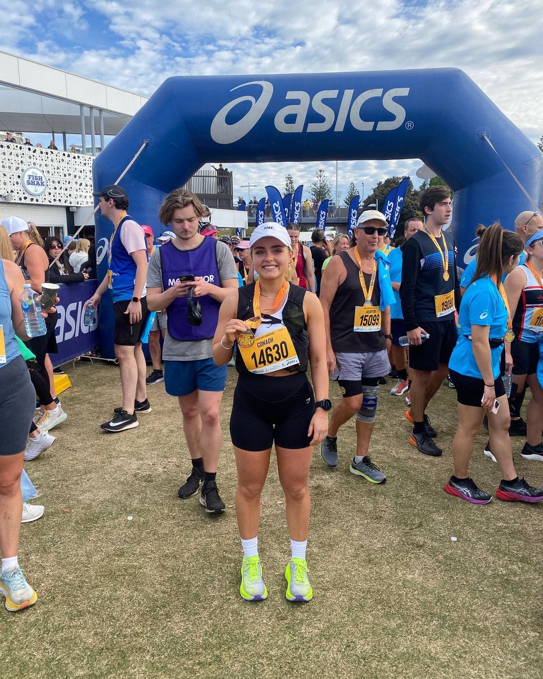 Female marathon runner in athletic wear with race number poses with medal after finishing race outdoors with crowd in background.