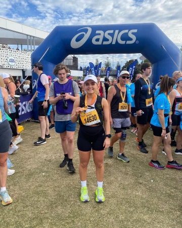 Female marathon runner in athletic wear with race number poses with medal after finishing race outdoors with crowd in background.