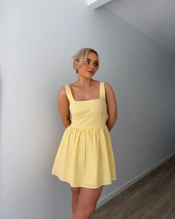 Blonde woman wearing a yellow minidress posing against an off-white wall indoors