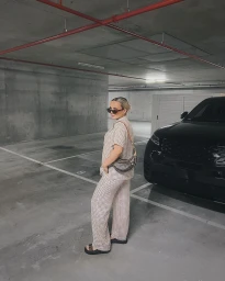 Woman in patterned short-sleeve shirt and matching trousers standing in an underground parking garage next to a black car.