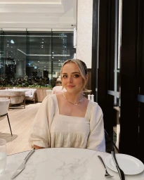Woman seated at a restaurant table with a white marble top, wearing a cream square-neck top and a silver necklace.