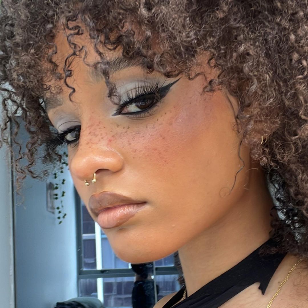 Close-up of a woman with curly hair and freckles, wearing makeup with dramatic eyeliner and lip gloss, posing indoors near a window.