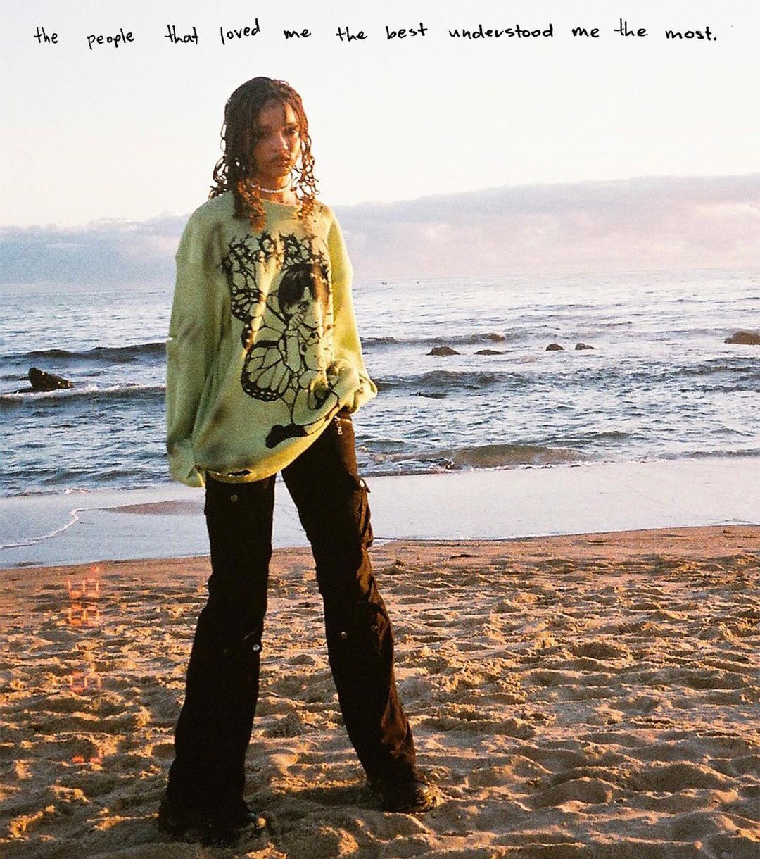 Young woman standing on beach at sunset with waves and sky. She is wearing a graphic sweatshirt and pants with ocean in the background.