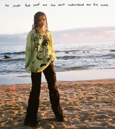 Young woman standing on a sandy beach at sunset wearing a light green graphic sweatshirt and black cargo pants