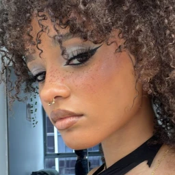 Close up portrait of a young woman with curly hair, winged eyeliner, freckles, and a gold septum piercing by Crystal Russell