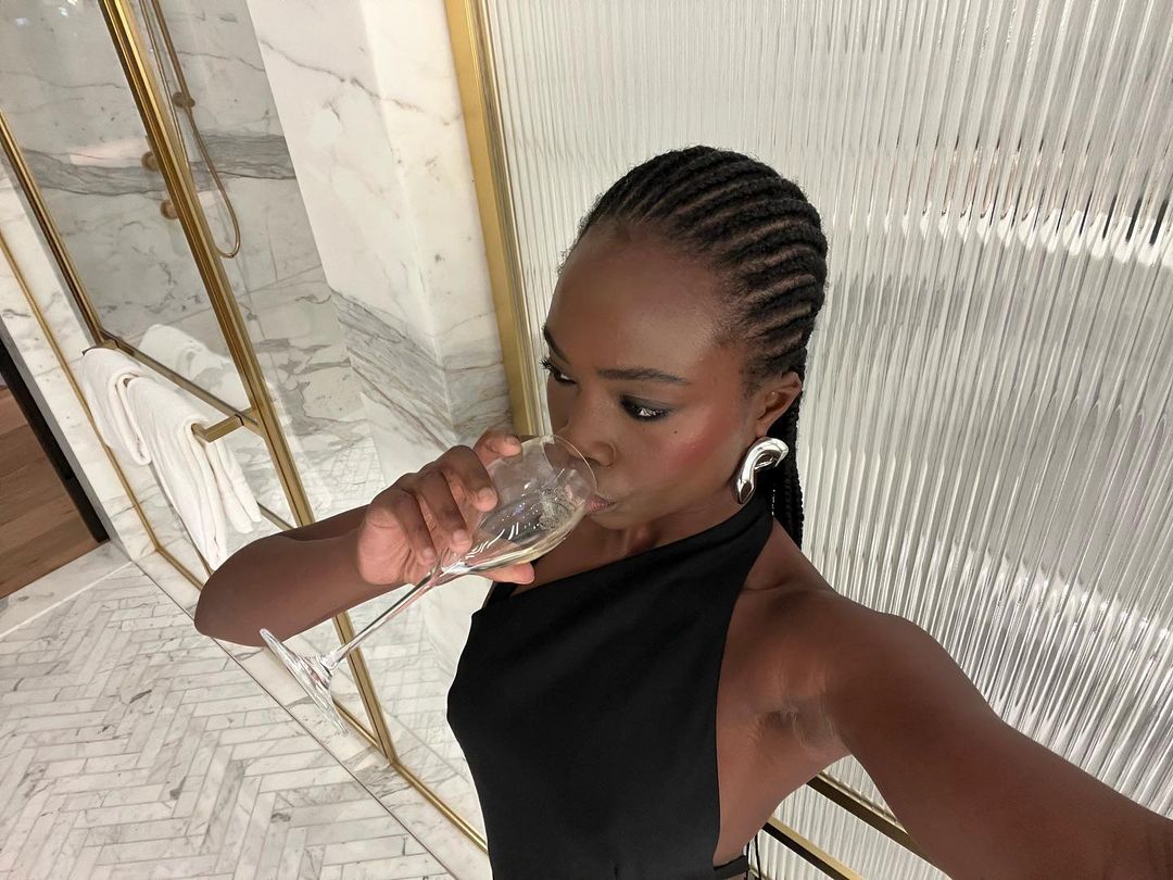 Woman in black dress drinks from a champagne glass indoors with modern gold and white decor, silver earrings, and braided hairstyle.
