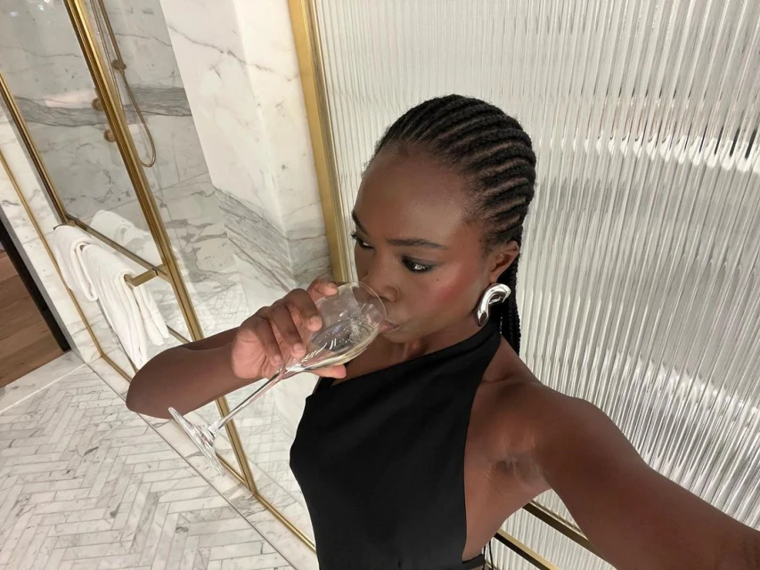 Woman in black dress drinks from a champagne glass indoors with modern gold and white decor, silver earrings, and braided hairstyle.