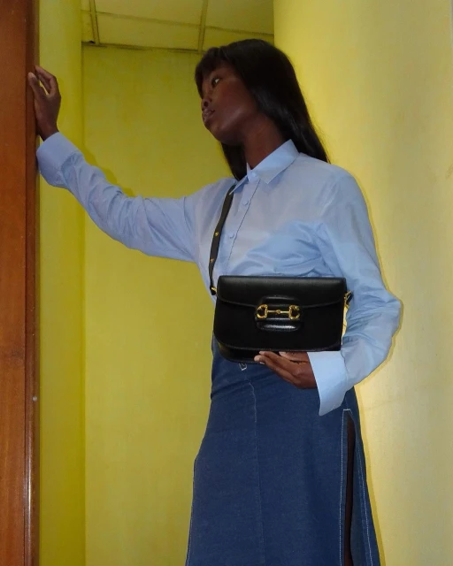 Adult female wearing a blue button-down shirt and denim skirt with a black purse, photographed indoors against a yellow wall.