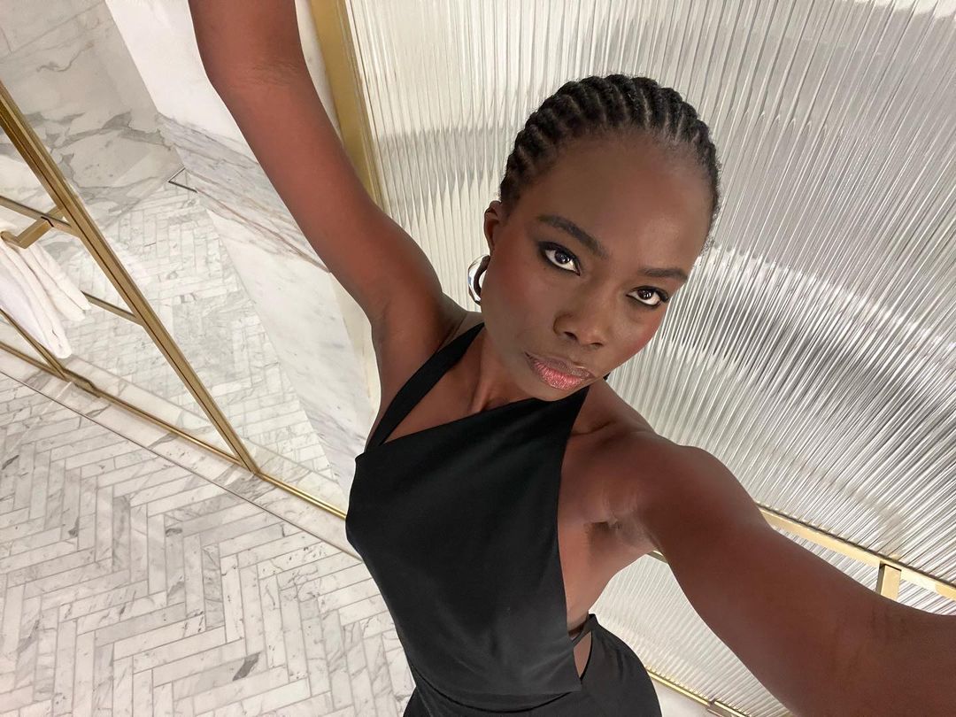 Portrait of an adult woman with braided hair in a black dress, posing in a white and gold bathroom setting, looking directly at the camera.