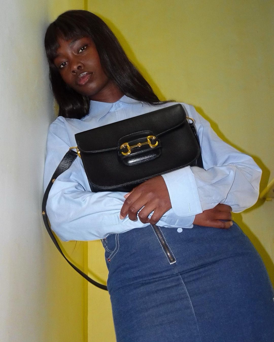 A Black woman in a blue button-down shirt and denim skirt poses with a black purse with gold hardware against a yellow wall.