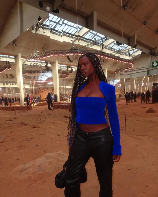 Fashionable Black woman in a bright blue top and black pants poses in a spacious, high-ceiling fashion show venue, exuding style.